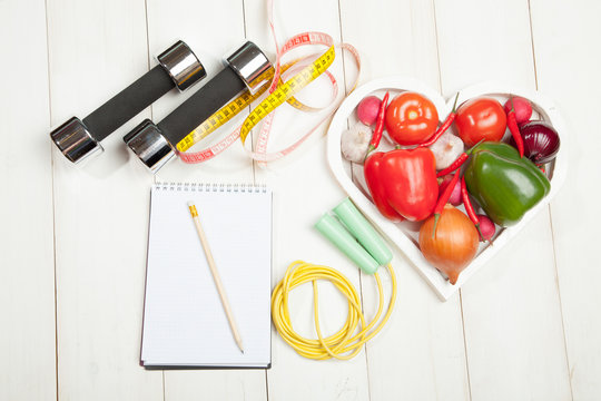 Sport and diet. Healthy lifestyle. Vegetables, dumbbells  notebook. Peppers, tomatoes, garlic, onion  radish in a heart on  white background