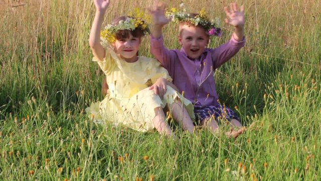 Happy little boy and girl in wreaths sit in dry grass and wave hands at summer evening