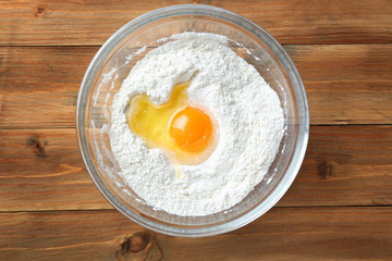 Glass bowl with flour and egg on wooden background