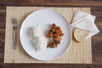 Scene with snow white ceramic dish of rice and roast meat, stew, meats with vegetables on a bamboo napkin on a wooden table from the old boards. Slices of white bread on the white paper napkins