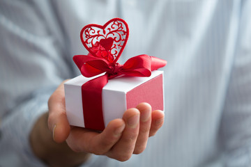 Close up shot man hand holding gift wrapped with red ribbon and heart. Small present in hands of human indoor. Selective focus on box. Holiday concept