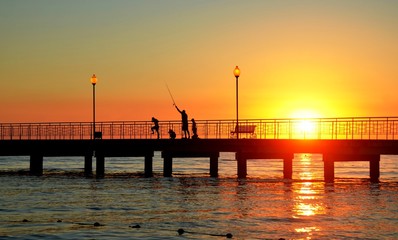 Young father with three young sons, rod catches fish in sea from pier at sunset. Backlight, soft focus, blur effect