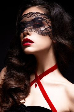 Young girl with a black lace mask on her face and a pendant in the form of heart on the neck. Beautiful model with bright make-up, red lips and curls. The picture on Valentine's Day.