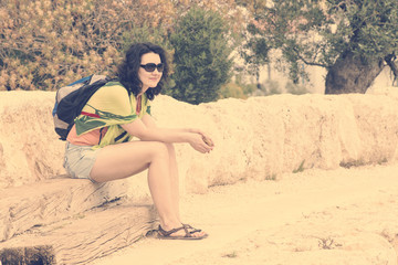 Woman sitting on wooden boards near a stone wall in sunglasses with a backpack