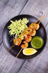 Cilantro grilled shrimps on skewers with sesame napa cabbage, green butter sauce and lemon. Skewered prawns on black plate. View from above, top studio shot