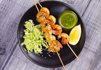 Cilantro grilled shrimps on skewers with sesame napa cabbage, green butter sauce and lemon. Skewered prawns on black plate. View from above, top studio shot