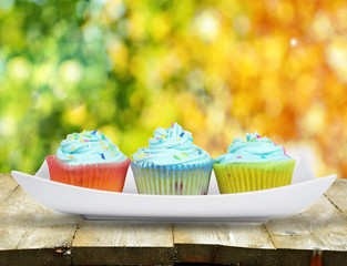 Three cupcakes with blue icing on plate on wooden table with bokeh of trees in the background