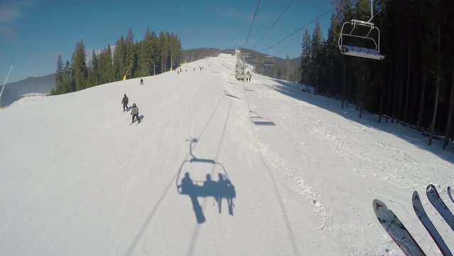 Ski lift moving up the mountain, with snow covered ski trails, skiers and snowboarders going downhill on a mountain slope below; first person pov shot; shadows on the snow below the ski lift chair