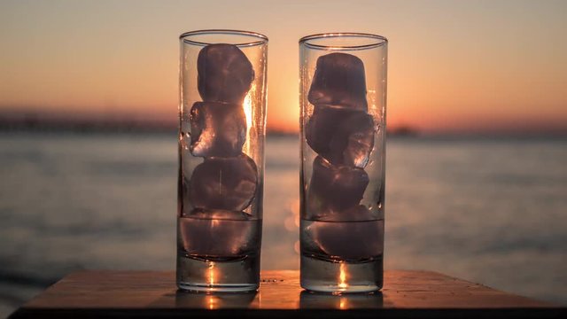 Timelapse close-up shot of two glasses with melting ice on the background of sea and sunset