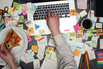 Top view on multitasking man working on laptop computer with post it notes all around his office desk. - obrazy, fototapety, plakaty