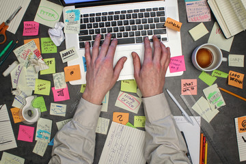 Top view on man working on laptop computer with post it notes all around his office table.