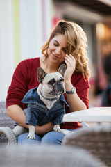 Beautiful woman holding adorable fawn french bulldog in her lap in cafeteria. 