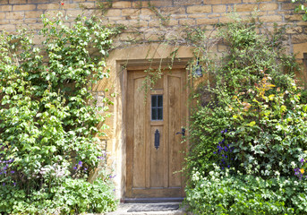 Fototapeta premium Light brown wooden doors in an old traditional English lime stone cottage surrounded by flowers in the front garden
