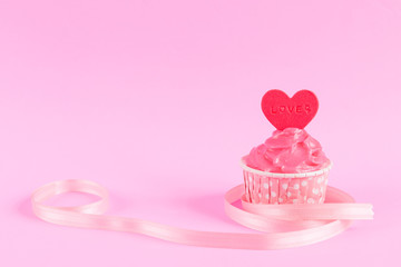 Sweet homemade cupcake with red heart shaped fondant over pink background with ribbon and copy...