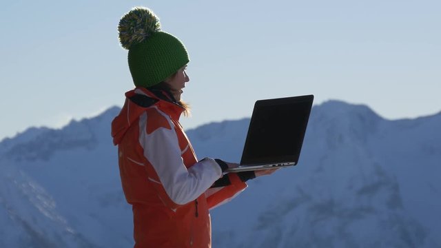 Woman dressed in winter clothes with cap talking on the network in the mountains with the help of a laptop