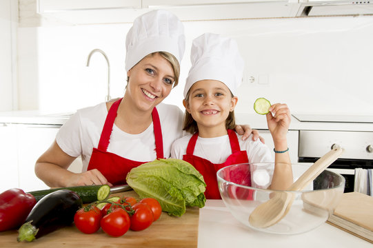 mother and little daughter cooking together with hat apron preparing salad at home kitchen