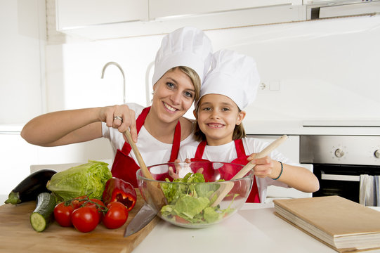 mother and little daughter cooking together with hat apron preparing salad at home kitchen