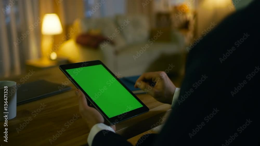 Poster at home man sits at his desk and uses tablet computer with green screen on it. his apartment is done