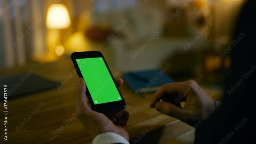 Poster at home man sits at his desk and uses smartphone with green screen on it. he drinks from a cup. his 