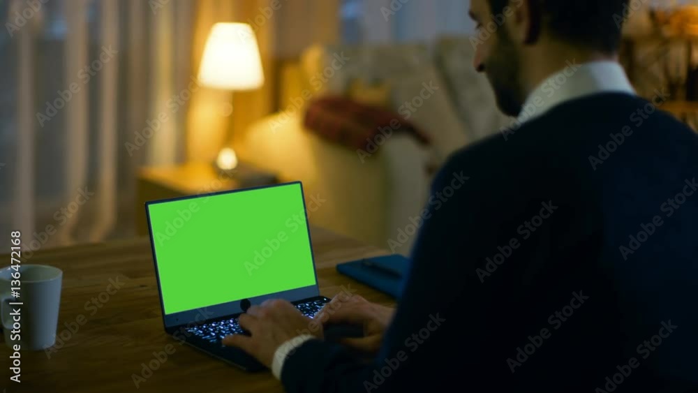 Poster at home back view of a man sitting at his desk and typing on a laptop with green screen on it. his a