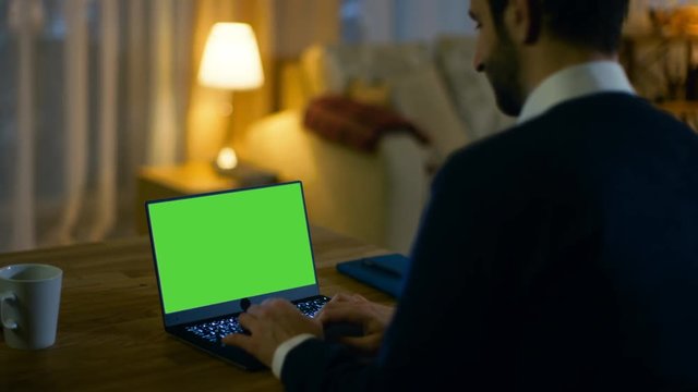 At Home Back View of a  Man Sitting at His Desk and Typing on a Laptop with Green Screen on It. His Apartment is Done in Yellow colours and is Warm. Shot on RED EPIC-W 8K Helium Cinema Camera.