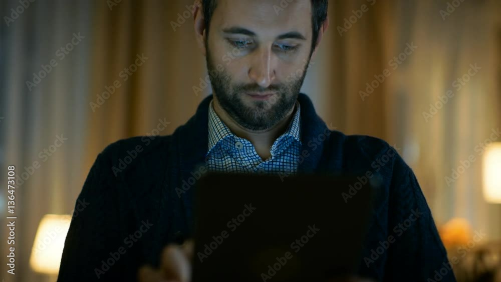 Poster portrait of a man at home holding and using tablet computer. screen illuminates him. his apartment i