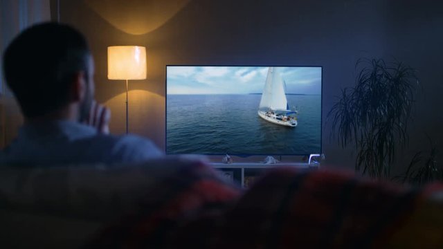 Back View of a Man Sitting on a Couch Watching Romantic Movie with Yachting and Camping in it on His Big Screen TV. It's Evening. Shot on RED EPIC-W 8K Helium Cinema Camera.