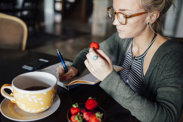 Woman with strawberry writing diary while sitting at table