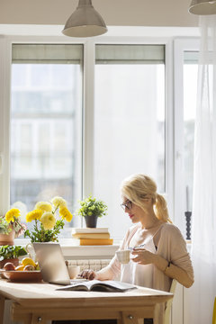 Woman with drink using laptop computer while sitting by window at home