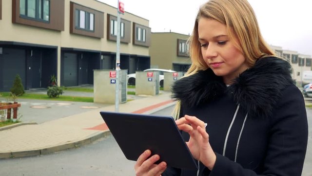 A young, beautiful woman stands on the street in a suburban area and works on a tablet