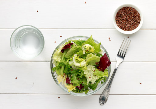 Mixed salad in glass bowl with flax seed and water on white wooden table.