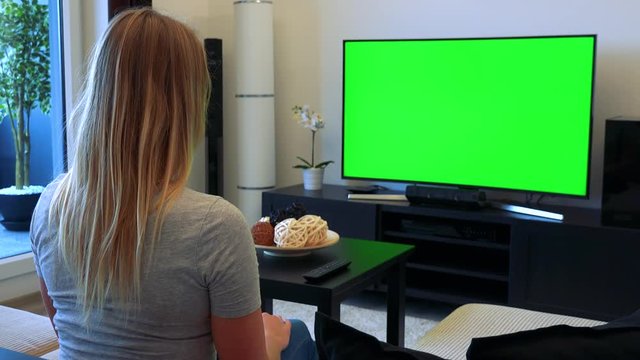 A young, beautiful woman sits on a couch in a living room and watches a TV with a green screen, then turns to the camera and shakes her head