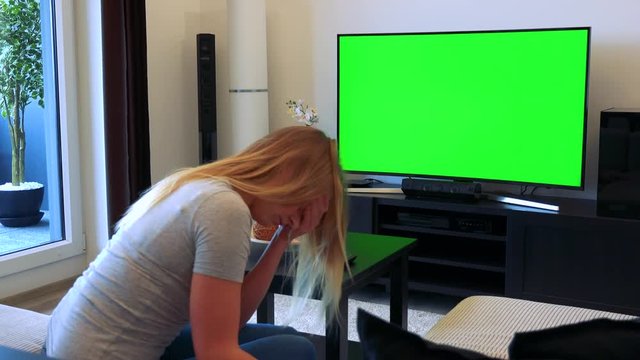 A blonde woman sits on a couch in a living room and watches a TV with a green screen, then covers her face with her hands unhappily
