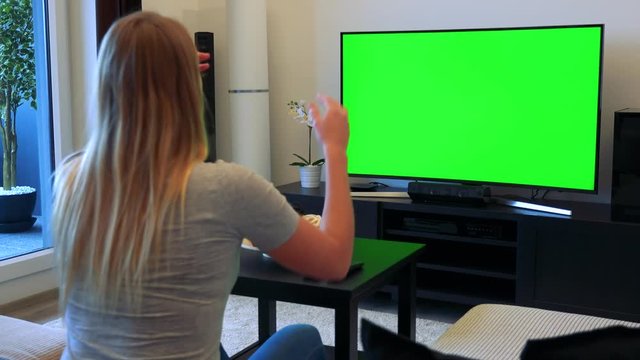 A blonde woman sits on a couch in front of a TV with a green screen in a living room, cheers and then celebrates