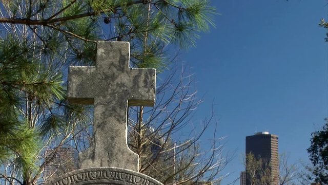 Camera pans down from large office buildings in the background to a cross shaped headstone.