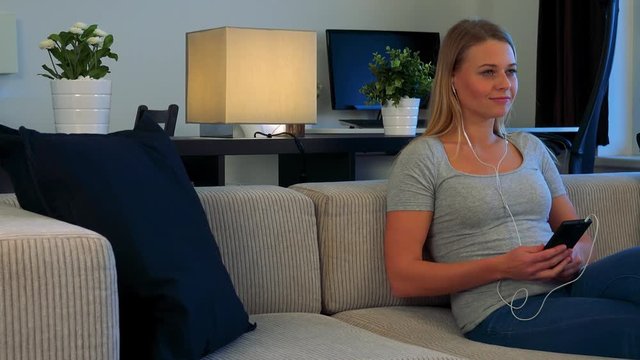 A young, beautiful woman sits on a couch in a living room and listens to music on a smartphonev
