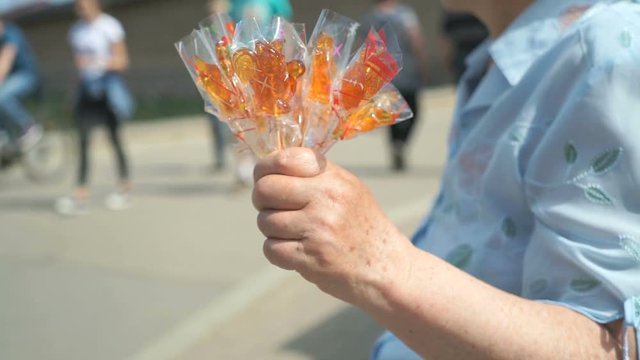 Elderly woman selling homemade sugar lollipops in individual packaging in summer street while passers going by sidewalk
