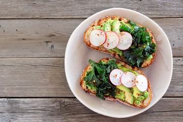 Plate of avocado toast with kale and radish on whole grain bread, above view over a rustic wood background