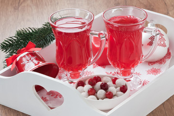 Cranberry hot drink / Cranberry hot winter drink in glass, berry sugar sweets on white wooden tray with candle, decorated ball and bell