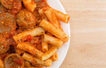 Top close view of rigatoni pasta with meatballs and sausage on a white plate atop a wood table.