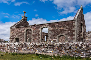 Fototapeta na wymiar Assynt Peninsula, Scotland - June 7, 2012: Lateral view on roofless ruins of Parliamentary Church with small cemetery and historic tomb stones. Brown-reddish stones. Blue sky with white clouds. Grass.