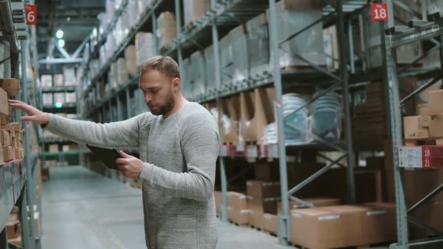 Young man using digital tablet in warehouse, checking goods list at supermarket. Touch the cardboard boxes. 4K