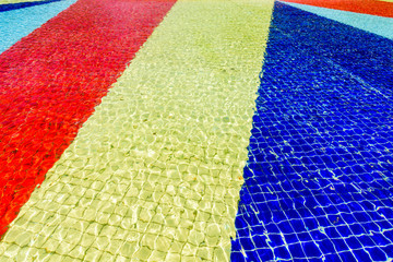 Light blue, red, yellow and blue striped mosaic floor of fountain covered with clear water at sunny day