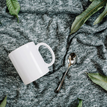 Blank template of white mug, spoon and green leaves on grey textile background. Flat lay, top view.