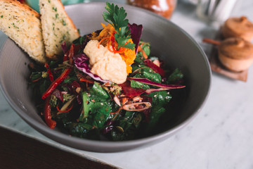 Close up og the dish with vegetarian salad with red pepper, cabbage, onion, lettuce, seeds and hummus on a marble table