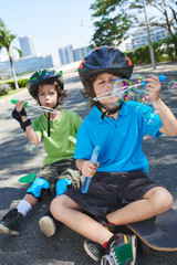 Fototapeta premium Handsome little brothers in colorful helmets sitting on skateboards and blowing soap bubbles in sunny park