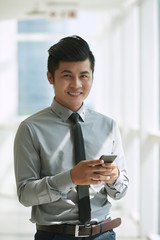 Cheerful Vietnamese businessman standing in office lobby while dialing number of his colleague on...