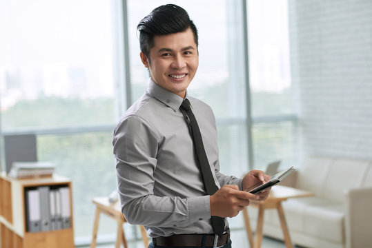 Waist-up portrait of smiling young businessman holding digital tablet in hands while standing in office lobby against panoramic window
