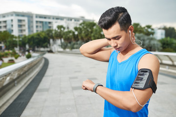 Waist-up portrait of pensive Asian athlete listening to music on smartphone and checking his progress with help of fitness tracker
