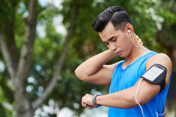 Portrait of pensive Asian athlete with stylish haircut rubbing his neck while looking at his fitness tracker in green park